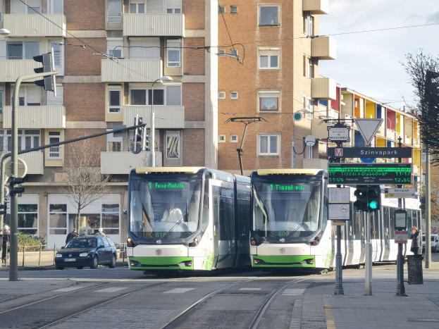Zwei grüne und weiße Straßenbahnen auf einer Stadtstraße mit hohen Gebäuden, Fahrzeugen, Fußgängern und städtischer Infrastruktur unter einem bewölkten Himmel.