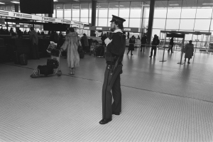 Polizeibeamter in Uniform mit Waffe, der in einem Flughafen-Terminal steht, umgeben von Menschen und Gepäck, mit Gepäckwagen, Deckenleuchten, Pfosten, Glaswänden und Informationsanzeigen im Hintergrund.