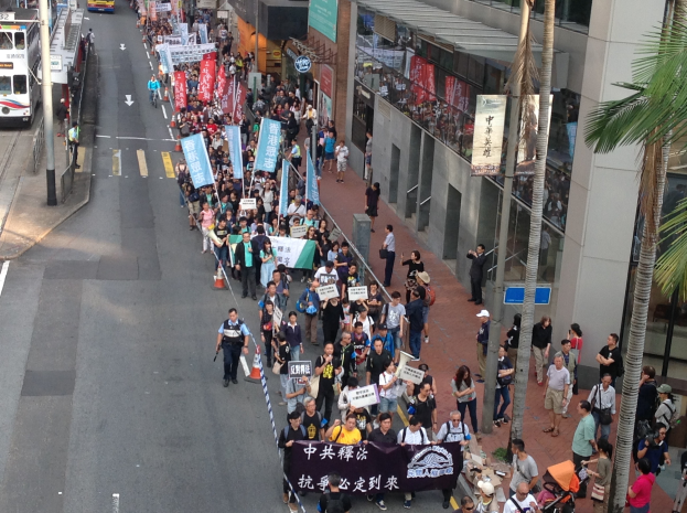 Großer Protestmarsch in Hong Kong mit Menschen mit Schildern und Plakaten, flankiert von Bäumen, Gebäuden, Fahrzeugen und Straßenschildern.
