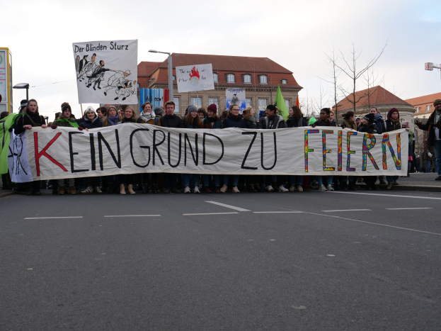 Eine Gruppe von Menschen hält ein Transparent mit der Aufschrift "Kein Grund zu Feiern" und protestiert gegen deutsche Sparmaßnahmen auf einer Stadtstraße mit Gebäuden, Bäumen und einem klaren Himmel.