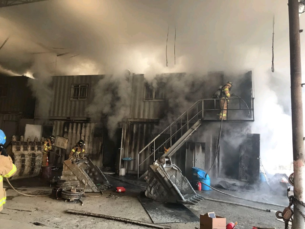 Feuerwehrleute in Helmen und Schutzausrüstung verwenden Schläuche, um ein Lagerfeuer zu löschen, mit Rauch, der aus dem Gebäude aufsteigt und Trümmern, die auf dem Boden verstreut sind.