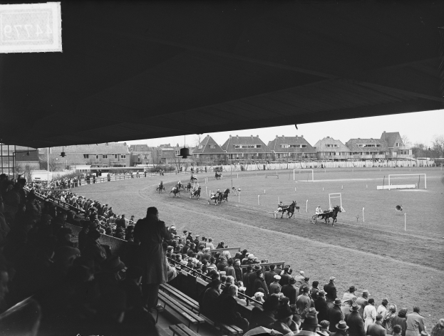 Schwarz-weiß-Foto eines Pferderennens in einer Stadion mit Zuschauern auf Bänken und Jockeys, die Pferde auf der Bahn reiten, mit Gebäuden, Bäumen, Pfählen und einem klaren Himmel im Hintergrund.