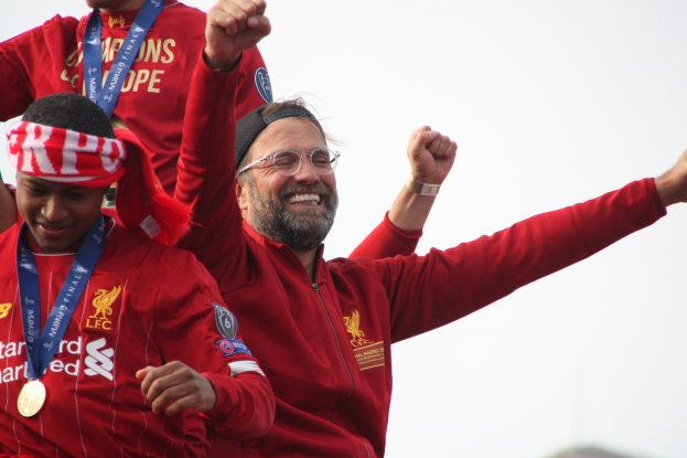 Liverpool-Trainer Jürgen Klopp feiert mit seinen Spielern nach dem Gewinn der UEFA Champions League, während zwei medalientragende Personen in der Nähe sind und der Himmel im Hintergrund zu sehen ist.