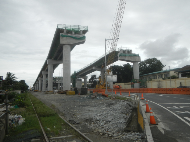 Baustelle mit einer Brücke im Hintergrund, Straße mit Verkehrskegeln markiert, verstreute Steine und Gras, eine Eisenbahnschiene auf der linken Seite, Bäume und Gebäude auf beiden Seiten der Straße und ein bewölkter Himmel.