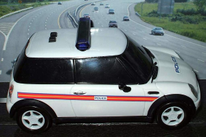 Ein weißer Polizeiwagen parkt vor einer belebten Straße mit fahrenden Fahrzeugen, umgeben von Gras und Bäumen in einer ländlichen Umgebung.