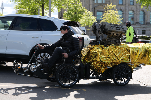 Ein Mann in einer schwarzen Jacke und Mütze sitzt in einem Rollstuhl mit einem großen Motor am Rücken, umgeben von Fahrzeugen auf einer Straße mit Bäumen, Gebäuden und Polen im Hintergrund unter einem klaren blauen Himmel.