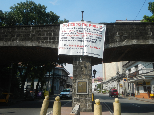 Verkehrsanzeigetafel an der Seite einer Brücke über einer Straße mit Fahrzeugen, mit Gebäuden, Bäumen, Strommasten und Drähten im Hintergrund unter einem klaren blauen Himmel.