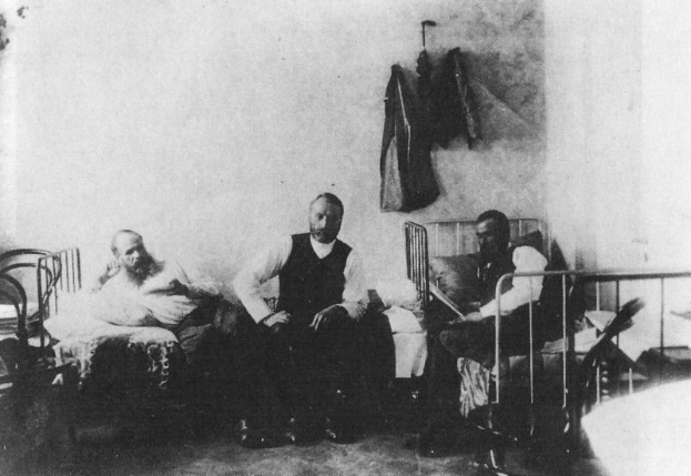 Schwarz-weiß-Foto von drei Männern, die in einem Krankenhauszimmer mit mehreren Betten, Stühlen und hängenden Körben sitzen, wobei ein Mann auf einem Bett ängstlich wirkt.