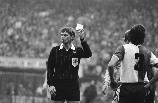 Ein Schiedsrichter hält eine rote Karte vor einer Menge von Menschen in Fußballtrikots, mit einem Zaun im Hintergrund, dargestellt in Schwarz-Weiß.