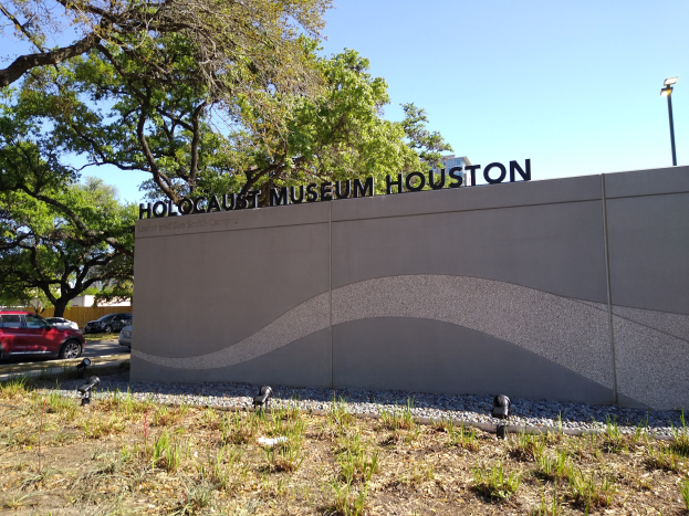 Außenansicht des Houston Holocaust Museums mit einer Gedenkwand, Steinen, Grünfläche, Bäumen, vorbeifahrenden Fahrzeugen, einem Straßenschild und einem bewölkten Himmel.