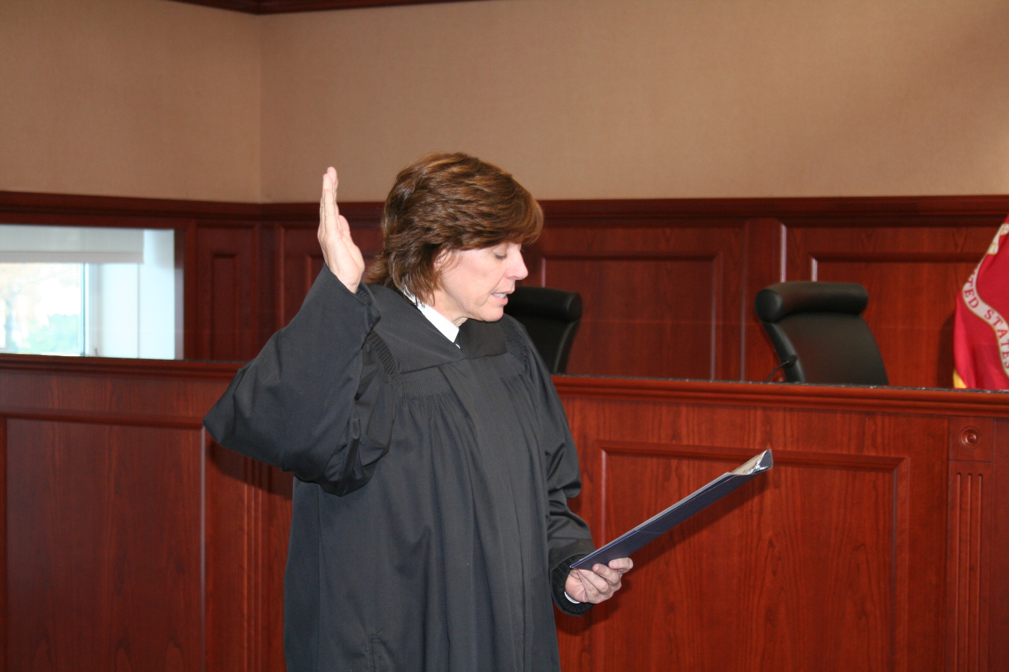 Eine Frau in einer schwarzen Robe steht an einem Richtertisch und hält eine Akte in der Hand, mit einem Mikrofon und einer Flagge auf dem Tisch, Stühle und ein Fenster im Hintergrund.