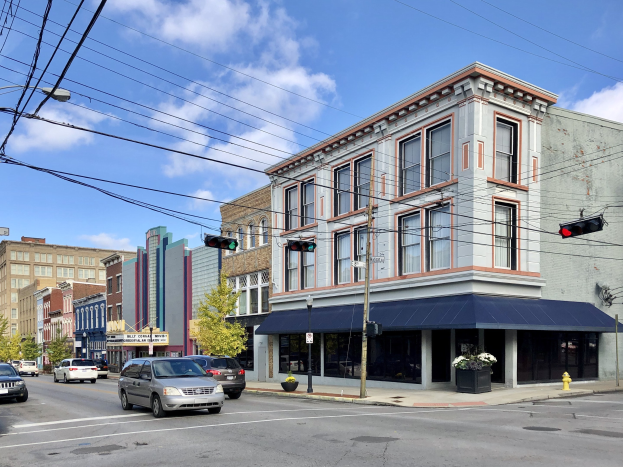 Eine Stra├če in einer Stadt mit parkenden Autos, Strommasten, Ampeln, Stra├čenlaternen, B├Ąumen und Geb├Ąuden unter einem bew├Âlktem Himmel, mit Text, der den Verkauf eines gewerblichen Grundst├╝cks f├╝r 1.250.000 Dollar ank├╝ndigt.