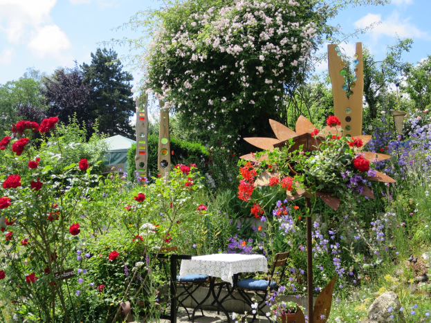Ein farbenfroher Garten auf der Chelsea Flower Show mit einem zentralen Tisch mit einer Decke, umgeben von bunten Blumen und Pflanzen, Bäumen im Hintergrund und einem bewölkten Himmel.