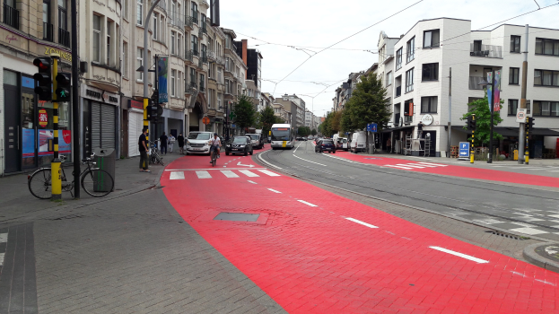 Rote Fahrradspur neben einer Stadtstraße mit Fahrzeugen, Fußgängern und Gebäuden auf beiden Seiten, Bäumen im Hintergrund unter einem klaren blauen Himmel.
