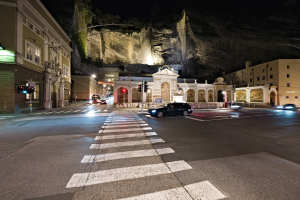 Eine Straße in einer Stadt bei Nacht mit fahrenden Autos, beleuchtet von Straßelaternen und Verkehrsampeln, Gebäuden mit Fenstern und Laternenmasten im Hintergrund und einem Hügel, mit einer Zebröquerung unten.