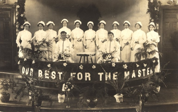 Eine Gruppe von Frauen in weißen Kleidern und Hauben steht auf einer Bühne mit einem Banner, auf dem 'Unser Bestes für den Meister' steht, umgeben von Blumentöpfen, Türen und einer Wand im Hintergrund.