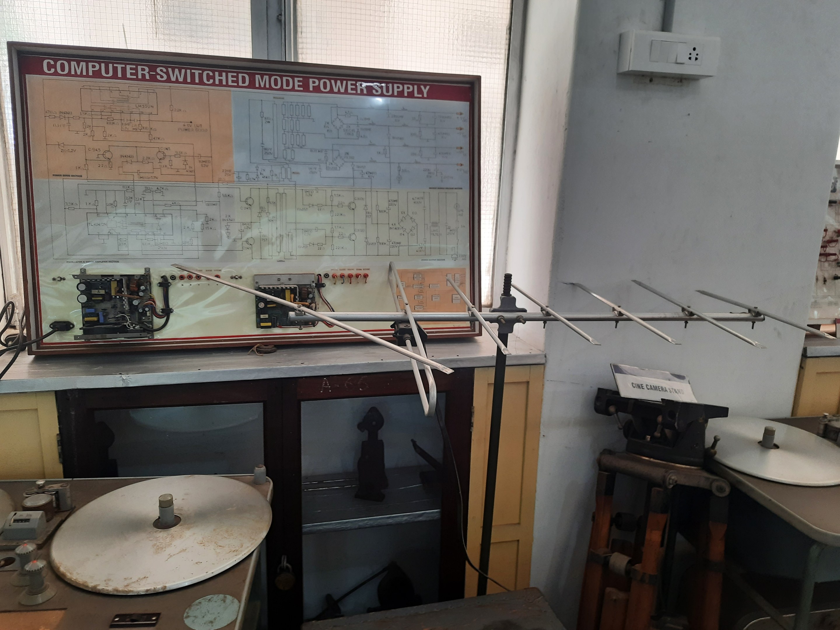 Ein unordentlicher Raum mit verschiedenen Elektronikgeräten, darunter eine beschriftete Leiterplatte auf einem Tisch, Metallstangen, eine wandmontierte Schalttafel, ein Fenster und Gegenstände in Schränken, die an eine computerge-switchte Mode-Leistungsversorgung erinnern.