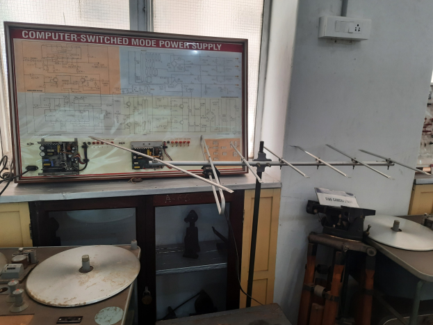 Ein unordentlicher Raum mit verschiedenen Elektronikgeräten, darunter eine beschriftete Leiterplatte auf einem Tisch, Metallstangen, eine wandmontierte Schalttafel, ein Fenster und Gegenstände in Schränken, die an eine computerge-switchte Mode-Leistungsversorgung erinnern.