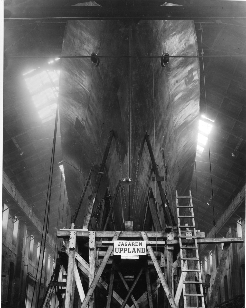Schwarzes und weißes Foto eines großen Bootes im Bau in einer Werft, das eine Leiter, hölzerne Pfosten, ein Schild, Metallrahmen, Deckenlampen und Fenster zeigt.