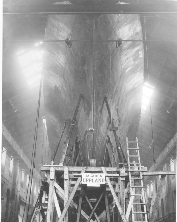 Schwarzes und weißes Foto eines großen Bootes im Bau in einer Werft, das eine Leiter, hölzerne Pfosten, ein Schild, Metallrahmen, Deckenlampen und Fenster zeigt.