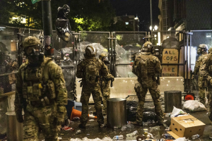 Eine Gruppe von Polizisten in Uniformen, Helmen und mit Taschen steht in einer Reihe vor einem Zaun bei Nacht, jeder hält eine Waffe in der Hand. Der Boden ist mit Mülltonnen, Flaschen, Kartons und anderem Schutt übersät. Im Hintergrund gibt es Bäume, Gebäude, Laternenpfähle und ein Schild an einem Pfahl.