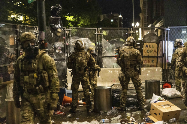 Eine Gruppe von Polizisten in Uniformen, Helmen und mit Taschen steht in einer Reihe vor einem Zaun bei Nacht, jeder hält eine Waffe in der Hand. Der Boden ist mit Mülltonnen, Flaschen, Kartons und anderem Schutt übersät. Im Hintergrund gibt es Bäume, Gebäude, Laternenpfähle und ein Schild an einem Pfahl.
