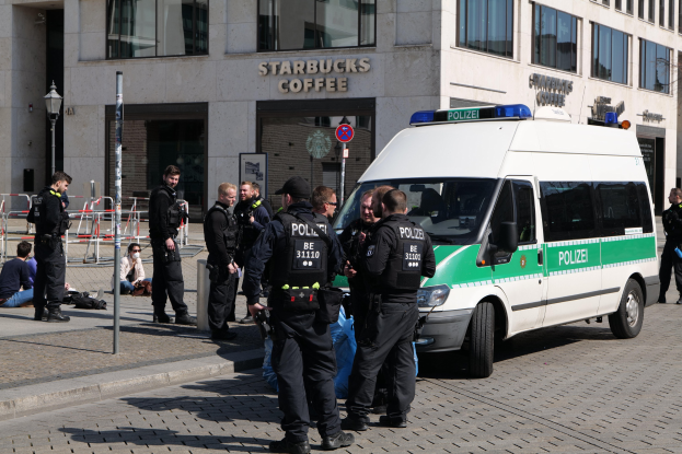 Eine Gruppe von Polizisten steht vor einem Starbucks-Café, rechts ein Van und links ein paar Menschen; im Hintergrund sind ein Gebäude, ein Schild, ein Laternenpfahl und ein Zaun zu sehen.