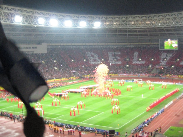 Ein Stadion mit Flutlicht, Eisengittern, Zuschauern, stehenden Personen, Werbetafeln, Bildschirmen, einer Überwachungskamera, Fahnen und Deko.