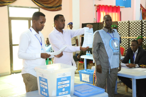 Eine Gruppe von Männern steht um einen Tisch mit Kisten und Papieren herum und gibt ihre Stimmen bei einer Wahlurne ab, mit Menschen, Tischen und verschiedenen Gegenständen im Hintergrund.