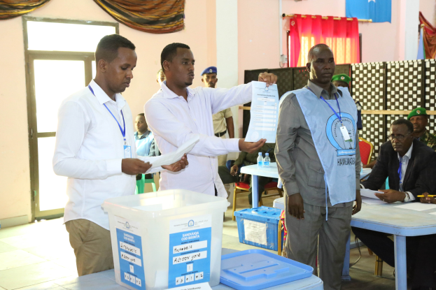 Eine Gruppe von Männern steht um einen Tisch mit Kisten und Papieren herum und gibt ihre Stimmen bei einer Wahlurne ab, mit Menschen, Tischen und verschiedenen Gegenständen im Hintergrund.