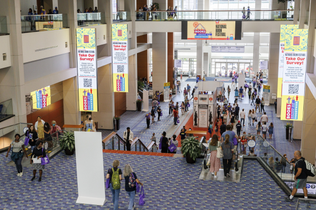 Eine große Menschenmenge bewegt sich durch ein großes Gebäude mit Bannern, Topfpflanzen, Rolltreppen, Geländern, Säulen, bedruckten Tafeln und Glastüren, einige tragen Taschen, was auf die Teilnahme am Nationalen Buchfest hinweist.