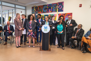Eine Gruppe von Menschen, die vor einem Rednerpult mit Mikrofon und einer Tafel mit der Aufschrift "Sancho-Stadtrat tritt dem Stadtrat bei" stehen und sitzen, mit Flaggen, Feuerlöscher und Deckenleuchten an der Rückwand.