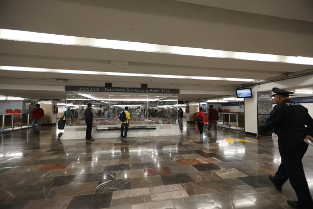 Ein belebter U-Bahnhof mit einem Polizeibeamten im Vordergrund, Menschen, die gehen, Anzeigetafeln und Bildschirme an der Decke sowie verschiedene bauliche Elemente wie Geländer.