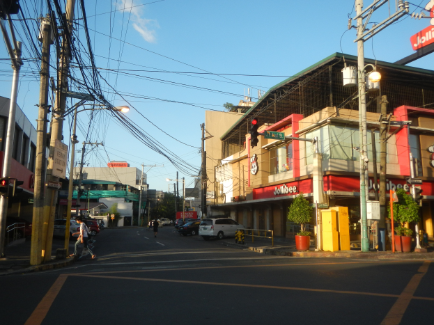 Eine belebte Stadtstraße mit Fußgängern, Fahrzeugen, Gebäuden und verschiedener städtischer Infrastruktur unter einem bewölkten Himmel.