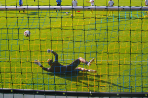 Ein Fußballspieler taucht ab, um einen Ball vor einem Tor zu halten, mit einem Netz im Vordergrund und ein paar Menschen im Hintergrund, sowie mehreren Fußballen auf dem Boden verstreut.