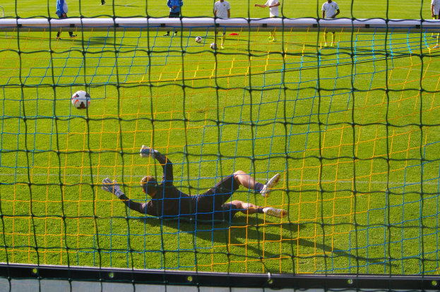 Ein Fußballspieler taucht ab, um einen Ball vor einem Tor zu halten, mit einem Netz im Vordergrund und ein paar Menschen im Hintergrund, sowie mehreren Fußballen auf dem Boden verstreut.