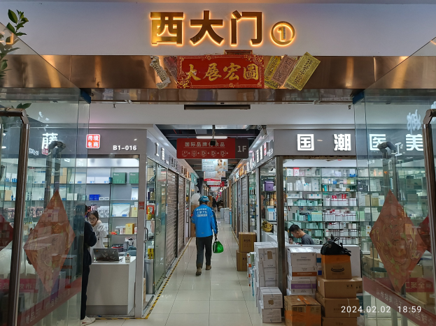 Innenraum einer chinesischen Apotheke mit Glaswänden, einer Pflanze links, Regalen voller Gegenstände und verstreuten Kartons sowie einem Namensschild oben.