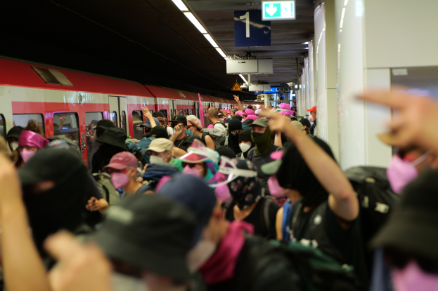 Eine Gruppe von Menschen mit Masken und Hüten steht vor einem Zug an einem Bahnhof; der Zug ist links und Säulen rechts; die Plattform hat eine Decke mit Lampen und Anzeigetafeln oben.