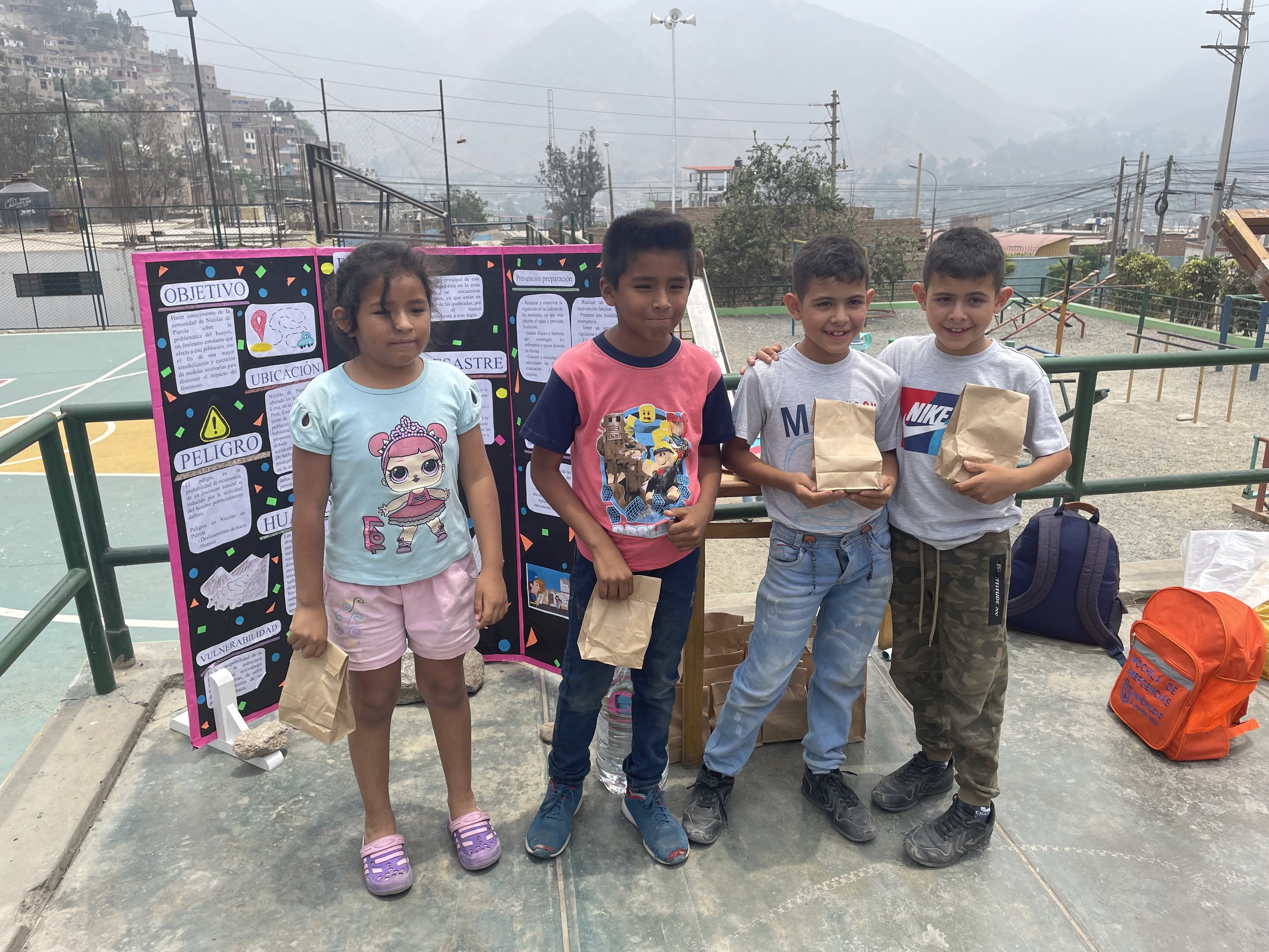 Gruppe von Kindern auf einem Basketballfeld mit Papiertüten, mit Materialien auf dem Boden und einer Tafel mit Text links daneben, vor einem Hintergrund aus Zaun, Pfählen, Straßenlaternen, Bäumen, Gebäuden, Hügeln und Himmel.