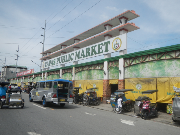Eine belebte Straßenkreuzung mit Autos, Motorrädern und Rikschas vor einem Gebäude, auf dem "Capas Public Market" steht, mit Strommasten, Laternenmasten, Gebäuden und einem bewölkten Himmel im Hintergrund.