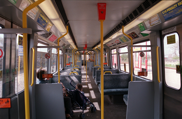 Innenansicht eines öffentlichen Verkehrsmittels mit Sitzreihen, einem Baby im Kinderwagen, offenen Fenstern und sichtbaren Hinweistafeln.