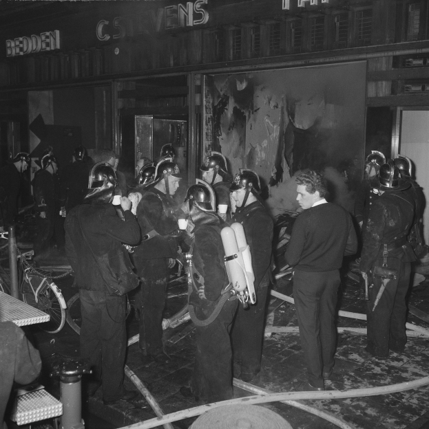 Schwarzes Foto von Feuerwehrleuten vor einem Gebäude, die Helme tragen und Sauerstoffflaschen tragen, mit einem Fahrrad und einem Tisch voller Gegenstände in der Nähe.