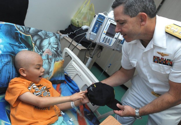 Ein Mann in weißer Uniform steht neben einem kleinen Jungen im Krankenhausbett, der eine Mütze in der Hand hält, mit medizinischen Geräten und Gegenständen im Hintergrund.