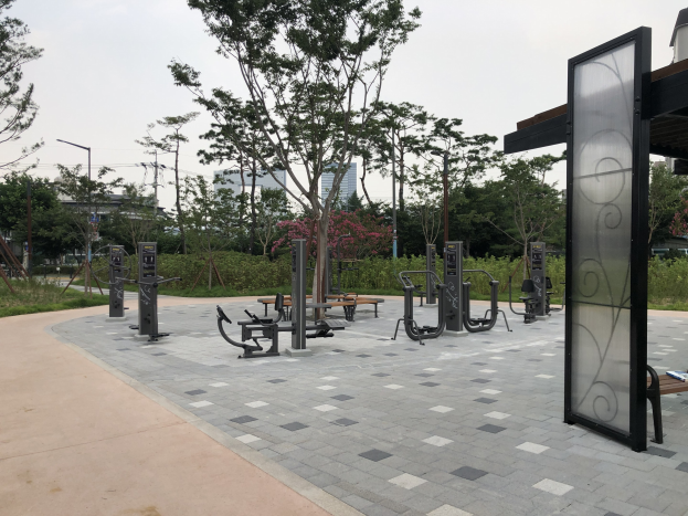 Outdoor-Park mit Fitnessgeräten, Bänken, Bäumen, Pflanzen, Gras, Pfählen, Lampen, Kabeln und Gebäuden vor einem Himmel-Hintergrund.