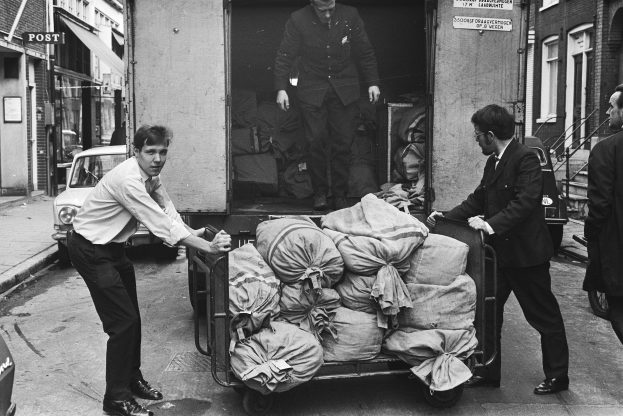 Eine Gruppe von Männern entlädt Taschen von einem geparkten Lastwagen am Straßenrand, mit einem vorbeifahrenden Auto und Gebäuden mit Fenstern, Treppen, Geländern und Texttafeln im Hintergrund, alles in Schwarz-Weiß dargestellt.
