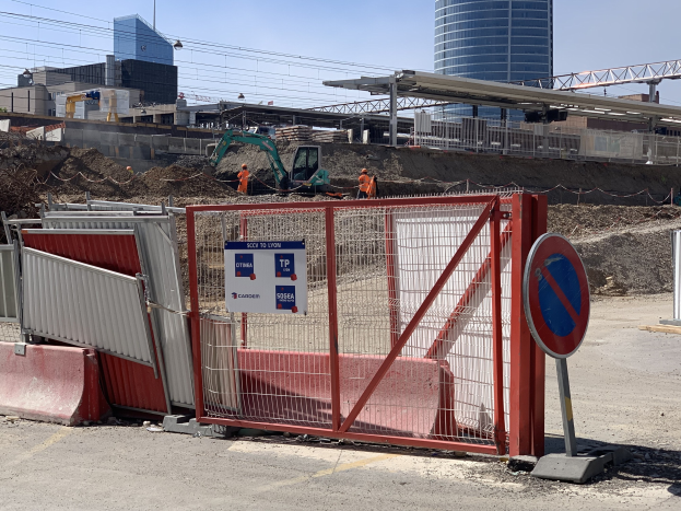 Baustelle mit rotem Tor und Schild, Metallzaun, Gebäuden, Kränen, Polen, Drähten, blauem Himmel, Arbeitern und Texttafeln im Hintergrund.