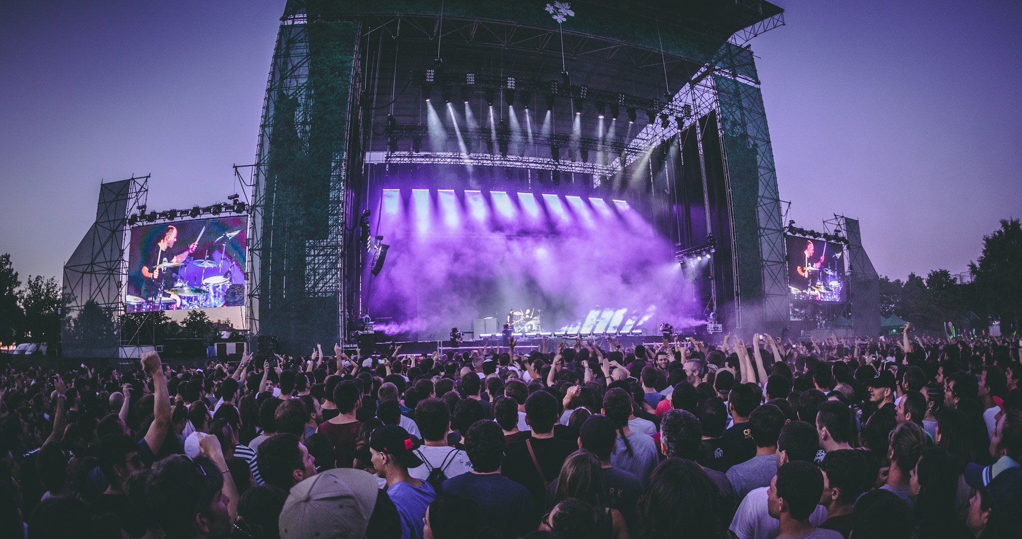 Eine große Menge bei einem Musikfestival schaut auf der hell erleuchteten Bühne mit Instrumenten und Bildschirmen, die vor einem Gebäude, Bäumen und einem klaren blauen Himmel aufgebaut sind.