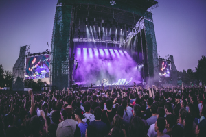 Eine große Menge bei einem Musikfestival schaut auf der hell erleuchteten Bühne mit Instrumenten und Bildschirmen, die vor einem Gebäude, Bäumen und einem klaren blauen Himmel aufgebaut sind.