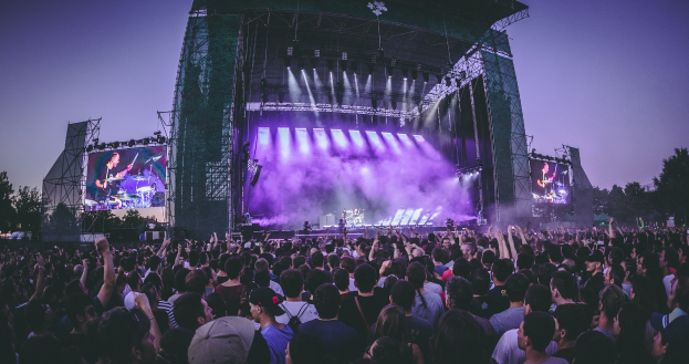 Eine große Menge bei einem Musikfestival schaut auf der hell erleuchteten Bühne mit Instrumenten und Bildschirmen, die vor einem Gebäude, Bäumen und einem klaren blauen Himmel aufgebaut sind.
