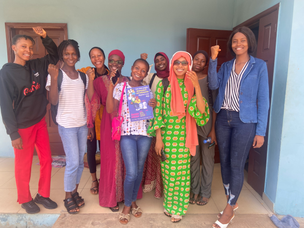 Eine Gruppe von Frauen steht nebeneinander vor einem Gebäude, lächelt und hält Bücher in den Händen, mit einem Tuch auf der rechten Seite und Türen und einer Wand im Hintergrund.